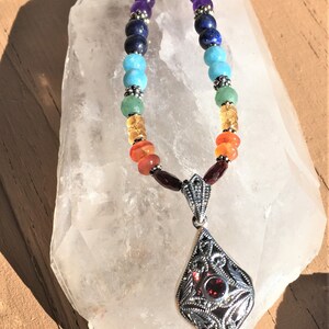 May include: A silver necklace with a gemstone pendant and a rainbow of colored beads. The beads are amethyst, lapis lazuli, turquoise, green aventurine, carnelian, and garnet. The pendant is a silver filigree design with a garnet stone in the center.