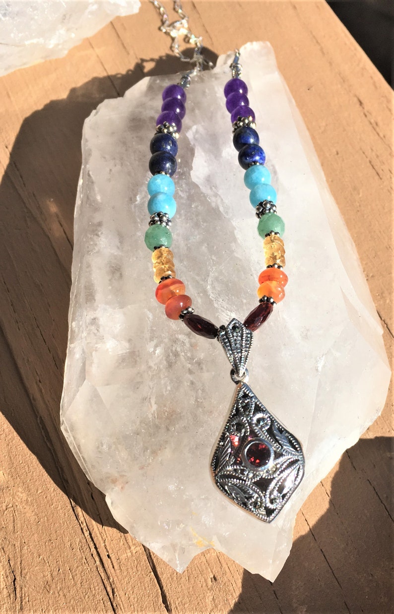 May include: A silver necklace with a gemstone pendant and a rainbow of colored beads. The beads are amethyst, lapis lazuli, turquoise, green aventurine, carnelian, and garnet. The pendant is a silver filigree design with a garnet stone in the center.
