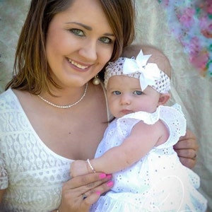 May include: A woman in a white lace dress holds a baby girl wearing a white dress and a white headband with a bow. The baby is looking at the camera.