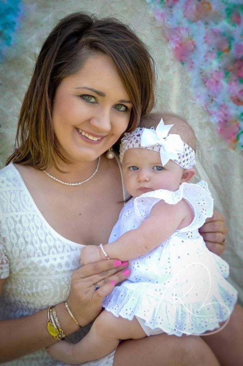 May include: A woman in a white lace dress holds a baby girl wearing a white dress and a white headband with a bow. The baby is looking at the camera.