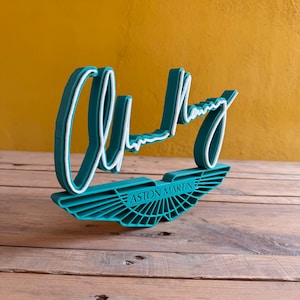 Può includere: Un cartello stampato in 3D di colore verde turchese con le parole "Aston Martin" e una firma bianca che recita "Alun Huyg"