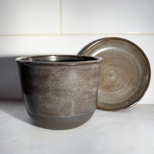 Może przedstawiać: Ceramiczna doniczka z nakrapianą szaro-brązową glazurą w górnej części i jednolitą ciemnobrązową podstawą, której towarzyszy pasujący okrągły spodek ustawiony pionowo za doniczką, oba elementy posiadają gładkie, szkliwione wykończenie.