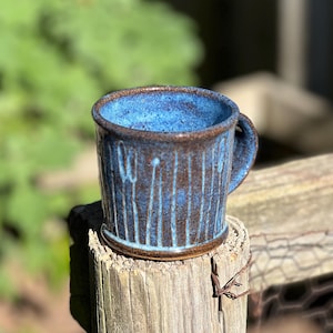 Puede incluir: Una taza de cerámica hecha a mano con un esmalte azul y líneas verticales texturizadas. La taza tiene un borde y un asa marrones. La taza mide aproximadamente 10 cm de alto.
