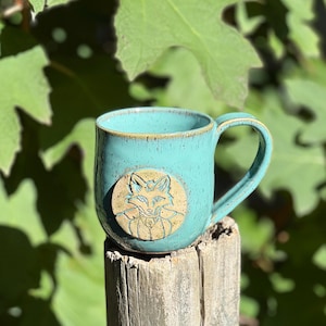 May include: A turquoise ceramic mug with a fox design. The mug has a round fox relief on the front and a curved handle. The mug is sitting on a wooden post, with green leaves in the background.