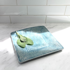 May include: A square, ceramic serving plate with a textured, wave-like pattern in shades of blue. Fresh green sage leaves rest on the plate. The plate is on a white marble surface.