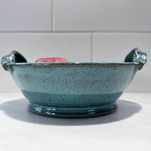 May include: A round, turquoise ceramic bowl with a speckled finish and two small handles on opposite sides. The bowl features a wide, shallow basin and a raised circular base. A single red apple is partially visible inside the bowl.