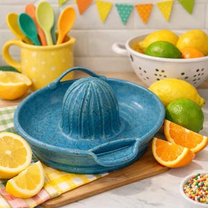 May include: A speckled turquoise ceramic citrus juicer with a handle, surrounded by lemons, limes, and orange slices. A yellow polka-dot mug with colorful spoons and a bowl of fruit are in the background.