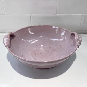 May include: A round, light lavender ceramic bowl with a glossy finish and dark speckles throughout. The bowl features two small, curved handles on opposite sides, a wide opening, and a smooth, rounded base, presenting a simple, functional design.
