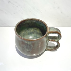 May include: A ceramic mug with a rounded body and a unique double-stacked handle design. The exterior and interior feature a mottled glaze in earthy tones of brown and sage green, with a smooth, glossy finish throughout the entire piece.