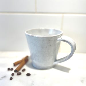 May include: A white ceramic mug with a speckled finish and a wide handle. The mug has a slightly flared rim and a tapered base. Beside the mug are two brown cinnamon sticks and several dark brown roasted coffee beans scattered on the surface.