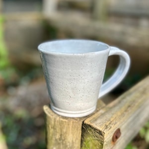 以下が含まれることがあります： 斑点のある釉薬が施された白いセラミック製のマグカップ。マグカップは丸みを帯びた形とハンドルが付いています。
