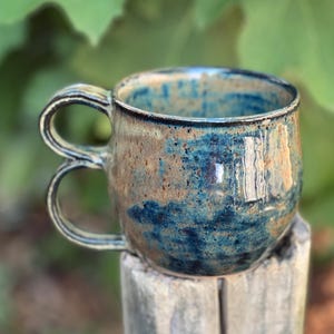 May include: Handmade ceramic mug with a unique double handle design. The mug features a blend of blue and brown glaze, creating a textured, earthy appearance. The mug is sitting on a wooden post, with a blurred green background.