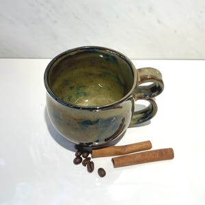 May include: A round ceramic mug with a glossy, mottled olive green and blue glaze, featuring a double-stacked handle. The mug is accompanied by several dark brown roasted coffee beans and two light brown cinnamon sticks placed in front.