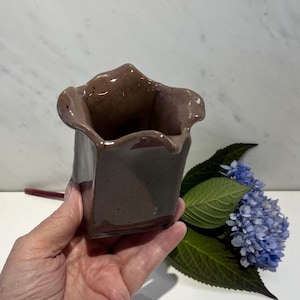 May include: A small, square, ceramic vase with a ruffled rim. The vase is a muted brown color with a glossy finish. A few blue hydrangea flowers and green leaves are next to the vase.
