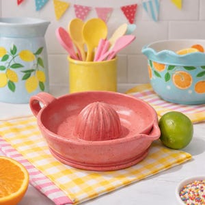 Puede incluir: Un exprimidor de cítricos de cerámica de color coral moteado con asa y escariador. El exprimidor está sobre un paño a cuadros amarillos y blancos. Otros utensilios de cocina incluyen una jarra azul con ilustraciones de limones, un soporte amarillo para utensilios y un cuenco turquesa.