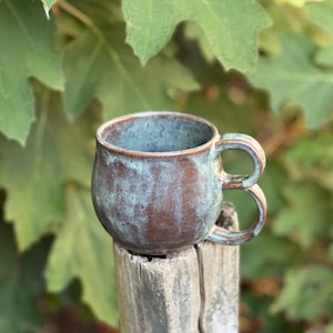 Puede incluir: Una taza de cerámica hecha a mano con un acabado texturizado único. La taza presenta un cuerpo redondeado y un asa distintiva. El color es una mezcla de tonos marrón y azul-verdoso, creando una estética rústica y terrosa. La taza mide aproximadamente 10 cm de alto.