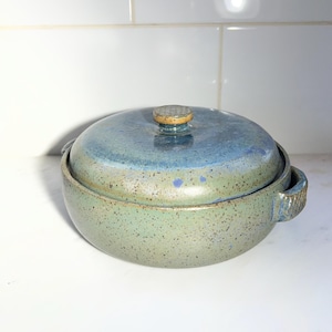 May include: A round ceramic lidded casserole dish with two side handles. The base is a speckled light green, while the lid features a gradient transition from green to blue. A small, textured circular knob sits atop the center of the lid.