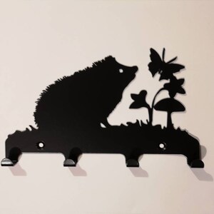 May include: Black metal key holder shaped like a hedgehog sniffing a flower with a butterfly. The key holder has three hooks.
