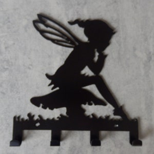 May include: Black metal key holder shaped like a fairy sitting on a mushroom. The fairy has wings and is holding a flower. The key holder has five hooks.