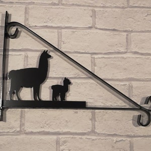 May include: Black metal wall-mounted shelf with a silhouette of two llamas. The larger llama is standing and the smaller llama is sitting. The shelf is triangular in shape and has a decorative scroll at the end.