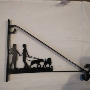 May include: Black metal wall mount with a silhouette of a couple walking two dogs. The mount is shaped like a triangle with a curved top and bottom.
