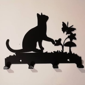 Puede incluir: Porta llaves de metal negro con una silueta de un gato que alcanza una mariposa y un hongo. El porta llaves tiene tres ganchos.