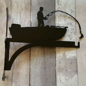 May include: Black metal silhouette of a person fishing from a boat. The silhouette is mounted on a black metal bracket. The bracket is designed to be attached to a wall.