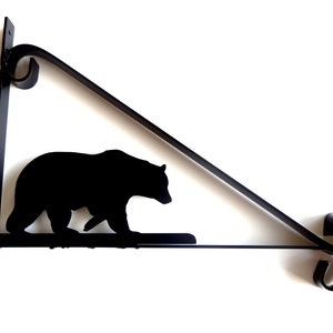 May include: Black metal hanging bracket with a bear silhouette. The bracket has a triangular design with decorative scrollwork. The bear is walking along a horizontal bar. The bracket is designed for hanging.