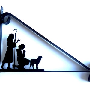 Op de afbeelding: Zwarte metalen silhouet van de kerststal met een herder, Maria, Jozef en een schaap. De silhouet is bevestigd aan een zwarte metalen beugel.