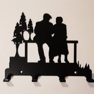 Könnte beinhalten: Schwarzer Schlüsselhalter aus Metall mit einer Silhouette eines Paares, das auf einer Bank mit Bäumen im Hintergrund sitzt. Der Schlüsselhalter hat drei Haken.