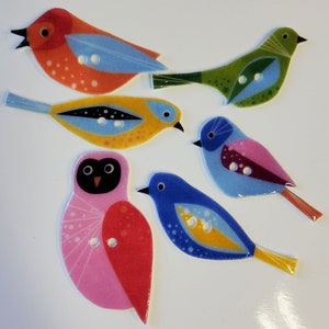 May include: A collection of six colorful bird-shaped buttons. The buttons feature various designs with shades of red, orange, yellow, green, blue, and pink. Each button has two holes for sewing and is set against a white background.