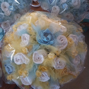 K&ouml;nnte beinhalten: Ein Babygeschenk-Bouquet, verpackt in Zellophan. Das Bouquet hat wei&szlig;e, blaue und gelbe Akzente, mit gerollten Artikeln, die Rosen &auml;hneln. Die Anordnung ist von gelbem Netz und kleinen floralen Verzierungen umgeben.