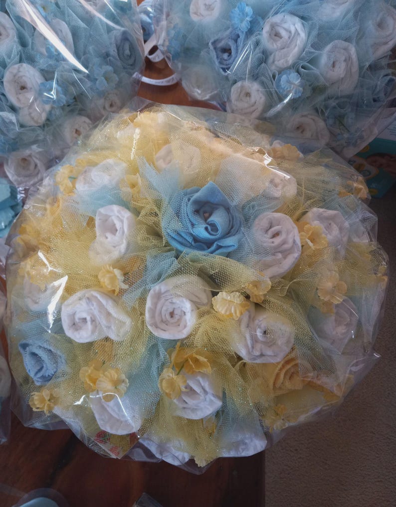 K&ouml;nnte beinhalten: Ein Babygeschenk-Bouquet, verpackt in Zellophan. Das Bouquet hat wei&szlig;e, blaue und gelbe Akzente, mit gerollten Artikeln, die Rosen &auml;hneln. Die Anordnung ist von gelbem Netz und kleinen floralen Verzierungen umgeben.