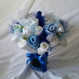 K&ouml;nnte beinhalten: Ein Geschenkstrau&szlig; zur Babyparty mit gerollten Windeln und Waschlappen, die wie Blumen angeordnet sind. Der Strau&szlig; enth&auml;lt wei&szlig;e, hellblaue und dunkelblaue Elemente, akzentuiert mit einem blauen Band und einem dekorativen Schmetterling. Die Anordnung wird in einem kleinen, transparenten Beh&auml;lter pr&auml;sentiert.
