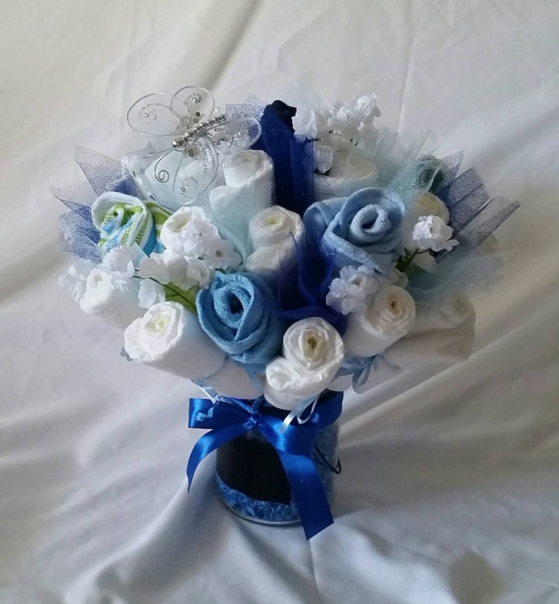 K&ouml;nnte beinhalten: Ein Geschenkstrau&szlig; zur Babyparty mit gerollten Windeln und Waschlappen, die wie Blumen angeordnet sind. Der Strau&szlig; enth&auml;lt wei&szlig;e, hellblaue und dunkelblaue Elemente, akzentuiert mit einem blauen Band und einem dekorativen Schmetterling. Die Anordnung wird in einem kleinen, transparenten Beh&auml;lter pr&auml;sentiert.
