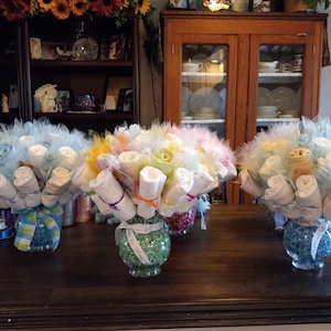 K&ouml;nnte beinhalten: Drei dekorative Tischdekorationen f&uuml;r eine Babyparty. Jedes Arrangement zeigt aufgerollte Babyartikel, die Blumen &auml;hneln, in Glasvasen. Die Str&auml;u&szlig;e sind mit T&uuml;ll und B&auml;ndern in Pastellfarben geschm&uuml;ckt.