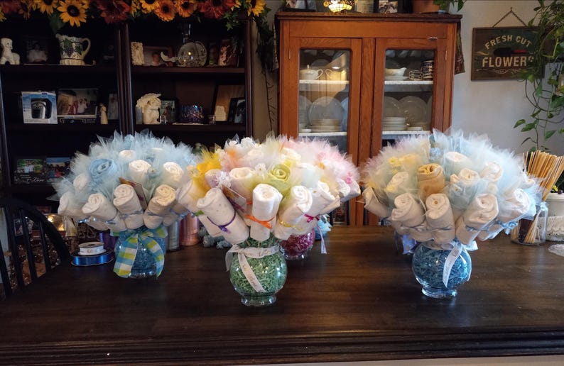 K&ouml;nnte beinhalten: Drei dekorative Tischdekorationen f&uuml;r eine Babyparty. Jedes Arrangement zeigt aufgerollte Babyartikel, die Blumen &auml;hneln, in Glasvasen. Die Str&auml;u&szlig;e sind mit T&uuml;ll und B&auml;ndern in Pastellfarben geschm&uuml;ckt.