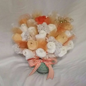 K&ouml;nnte beinhalten: Eine dekorative Anordnung, die einem Bouquet &auml;hnelt, mit gerollten wei&szlig;en und orangen Artikeln, m&ouml;glicherweise Windeln. Die Anordnung ist mit orangefarbenem T&uuml;ll, wei&szlig;en floralen Elementen und einer roten Blume akzentuiert. Ein gestreiftes orange-wei&szlig;es Band schm&uuml;ckt die Basis.