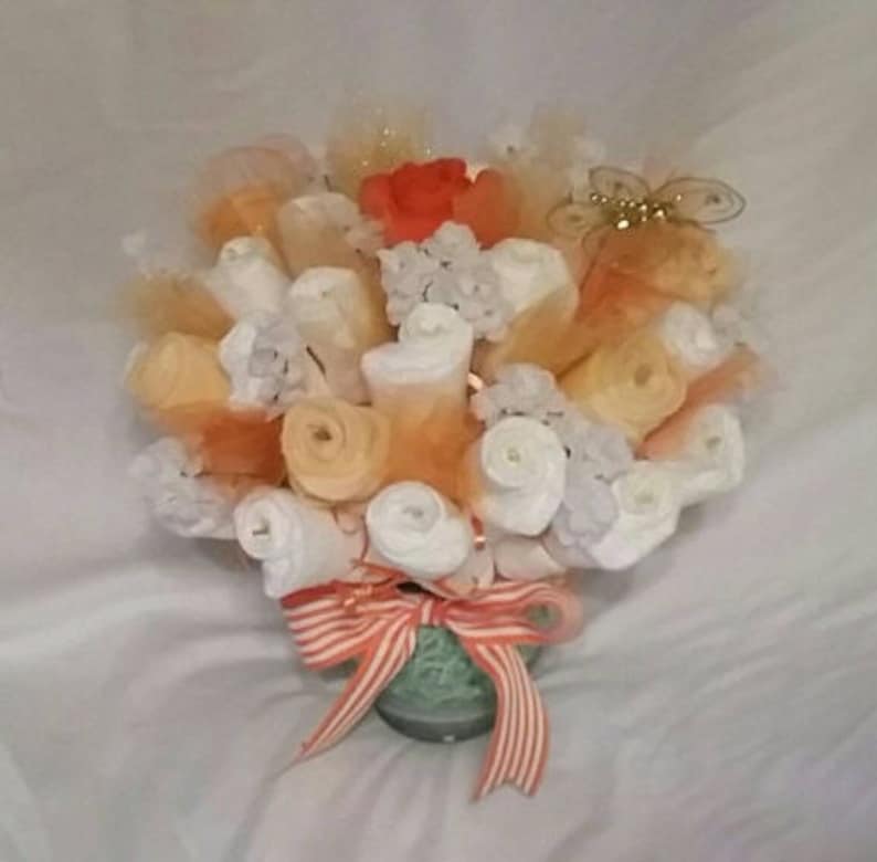 K&ouml;nnte beinhalten: Eine dekorative Anordnung, die einem Bouquet &auml;hnelt, mit gerollten wei&szlig;en und orangen Artikeln, m&ouml;glicherweise Windeln. Die Anordnung ist mit orangefarbenem T&uuml;ll, wei&szlig;en floralen Elementen und einer roten Blume akzentuiert. Ein gestreiftes orange-wei&szlig;es Band schm&uuml;ckt die Basis.