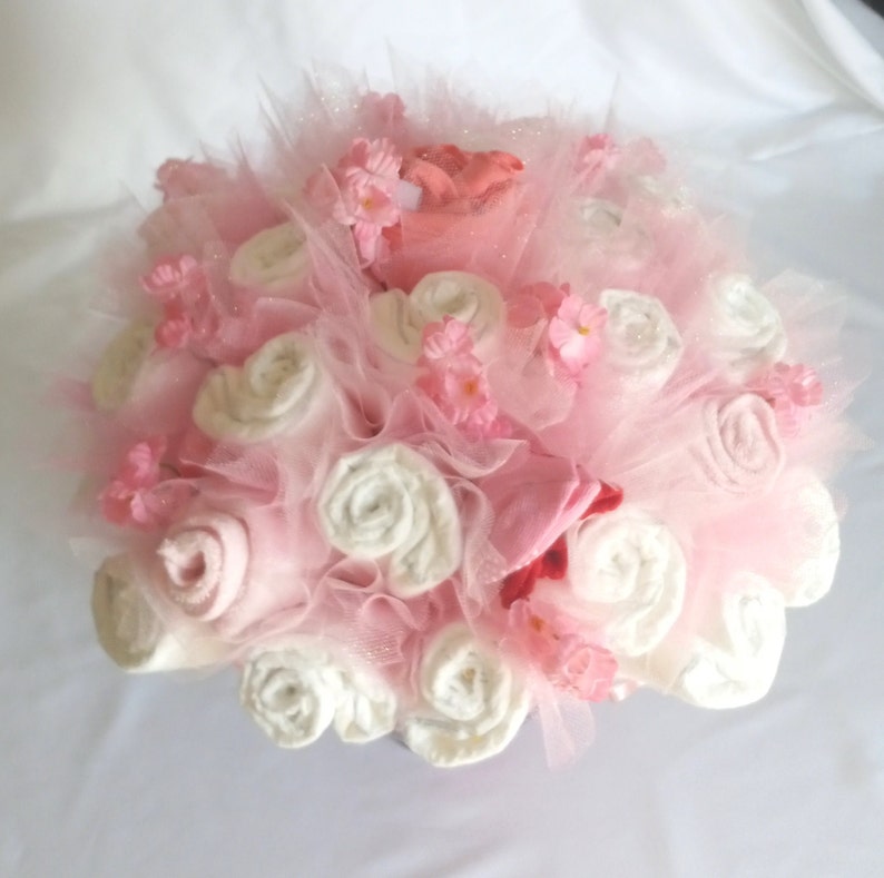 K&ouml;nnte beinhalten: Ein Geschenkarrangement f&uuml;r eine Babyparty mit gerollten wei&szlig;en und rosa Babyartikeln, die einem Blumenstrau&szlig; &auml;hneln. Die Anordnung ist mit rosa T&uuml;ll, kleinen rosa Bl&uuml;ten und einem Hauch von Rot verziert. Das Farbschema ist zartrosa und wei&szlig;.