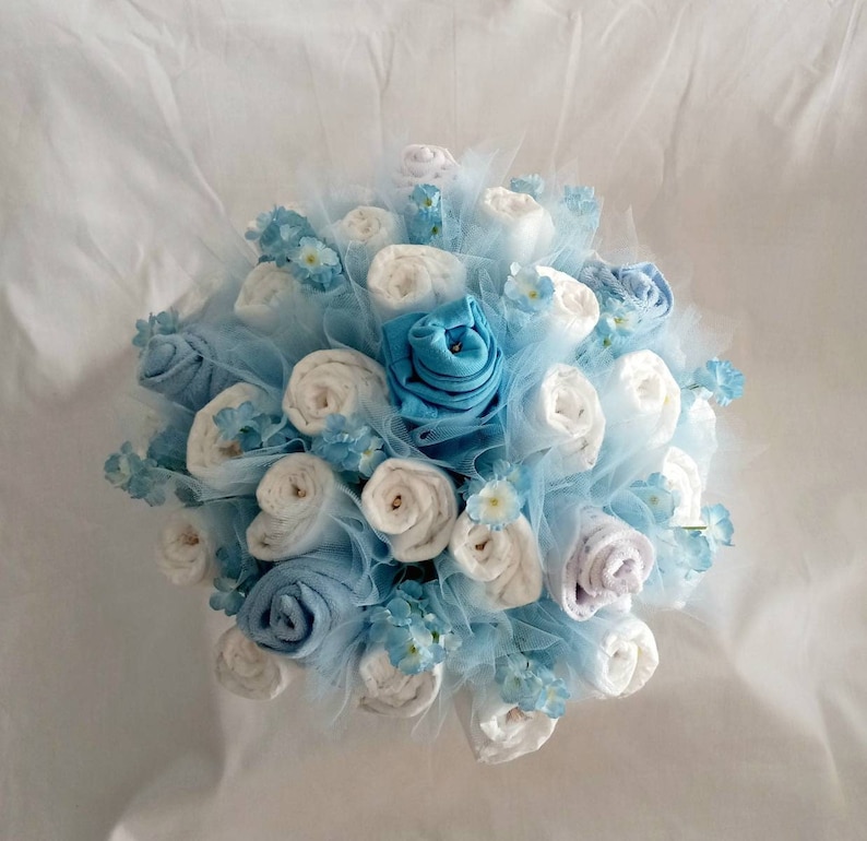 K&ouml;nnte beinhalten: Ein Bouquet aus gerollten wei&szlig;en und blauen Stoffartikeln, die Blumen &auml;hneln, ist auf einer hellblauen T&uuml;llbasis angeordnet. Kleine blaue Blumenakzente sind &uuml;ber die Anordnung verstreut und erzeugen ein weiches, zartes Aussehen.