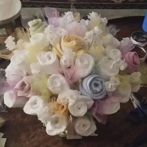 K&ouml;nnte beinhalten: Ein Babygeschenk-Bouquet in Pastellt&ouml;nen. Die Anordnung umfasst gerollte Baby-Waschlappen in Wei&szlig;, Gelb, Rosa und Blau, akzentuiert mit T&uuml;ll und kleinen wei&szlig;en Blumendekorationen. Das Bouquet wird auf einer dunklen Holzoberfl&auml;che pr&auml;sentiert.