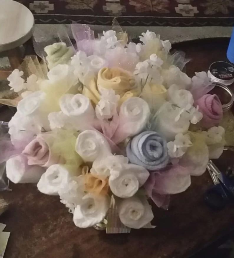 K&ouml;nnte beinhalten: Ein Babygeschenk-Bouquet in Pastellt&ouml;nen. Die Anordnung umfasst gerollte Baby-Waschlappen in Wei&szlig;, Gelb, Rosa und Blau, akzentuiert mit T&uuml;ll und kleinen wei&szlig;en Blumendekorationen. Das Bouquet wird auf einer dunklen Holzoberfl&auml;che pr&auml;sentiert.
