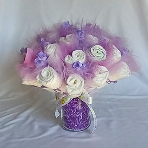 K&ouml;nnte beinhalten: Ein Babyparty-Mittelst&uuml;ck mit einem Bouquet aus gerollten Windeln, die Rosen &auml;hneln. Das Arrangement umfasst lavendelfarbenes T&uuml;ll, Federn und dekorative lila Akzente, pr&auml;sentiert in einem klaren Glasgef&auml;&szlig;, das mit lila zerkleinertem Papier gef&uuml;llt und mit einem Band gebunden ist.
