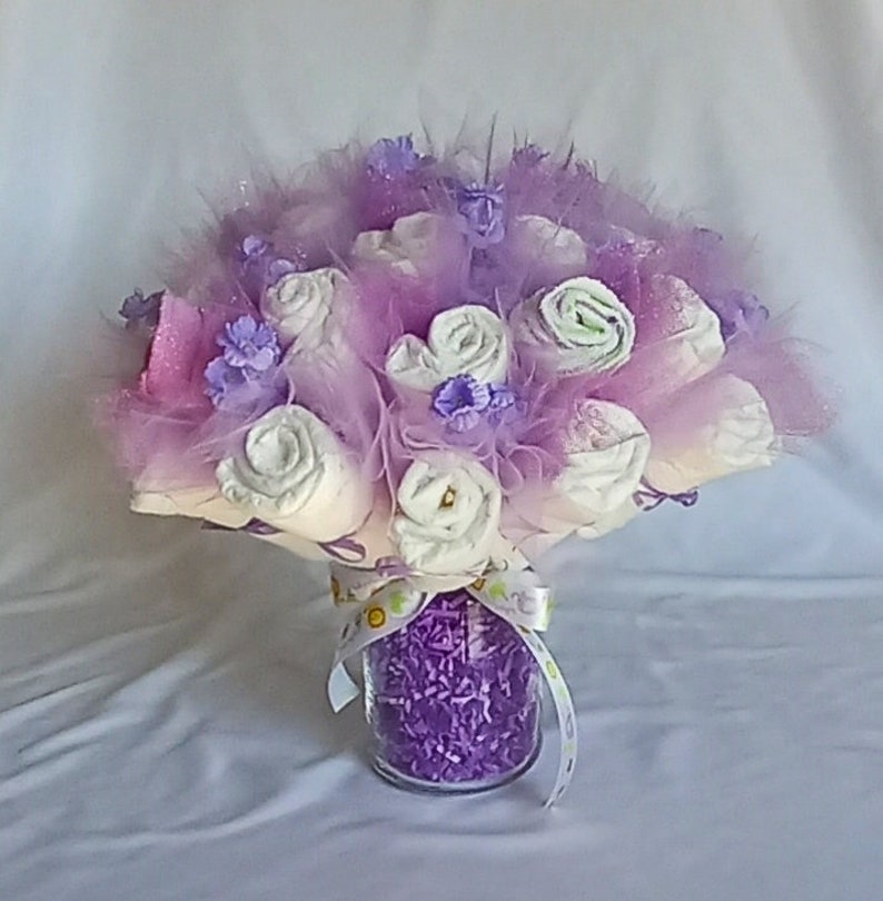 K&ouml;nnte beinhalten: Ein Babyparty-Mittelst&uuml;ck mit einem Bouquet aus gerollten Windeln, die Rosen &auml;hneln. Das Arrangement umfasst lavendelfarbenes T&uuml;ll, Federn und dekorative lila Akzente, pr&auml;sentiert in einem klaren Glasgef&auml;&szlig;, das mit lila zerkleinertem Papier gef&uuml;llt und mit einem Band gebunden ist.