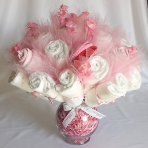 K&ouml;nnte beinhalten: Ein Geschenkarrangement f&uuml;r eine Babyparty in einer durchsichtigen Glasvase. Das Arrangement besteht aus gerollten wei&szlig;en Babywaschlappen, rosa T&uuml;ll und kleinen rosa Bl&uuml;ten. Ein rosa Babyhut ist in der Mitte eingebettet. Ein wei&szlig;es Band mit Text ist um die Vase gebunden.