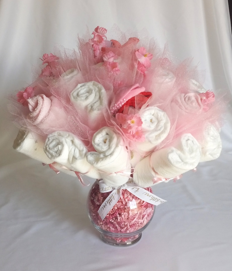 K&ouml;nnte beinhalten: Ein Geschenkarrangement f&uuml;r eine Babyparty in einer durchsichtigen Glasvase. Das Arrangement besteht aus gerollten wei&szlig;en Babywaschlappen, rosa T&uuml;ll und kleinen rosa Bl&uuml;ten. Ein rosa Babyhut ist in der Mitte eingebettet. Ein wei&szlig;es Band mit Text ist um die Vase gebunden.