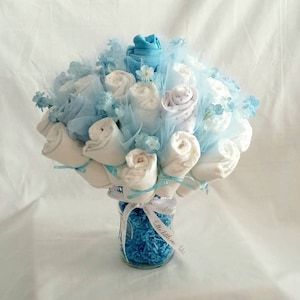 K&ouml;nnte beinhalten: Ein Babyparty-Geschenk, ein Strau&szlig; aus gerollten wei&szlig;en und blauen Babyartikeln, wie Socken und Waschlappen. Der Strau&szlig; ist in einem durchsichtigen Glasgef&auml;&szlig; angeordnet, das mit blauem Zierpapier gef&uuml;llt und mit einem Band gebunden ist.