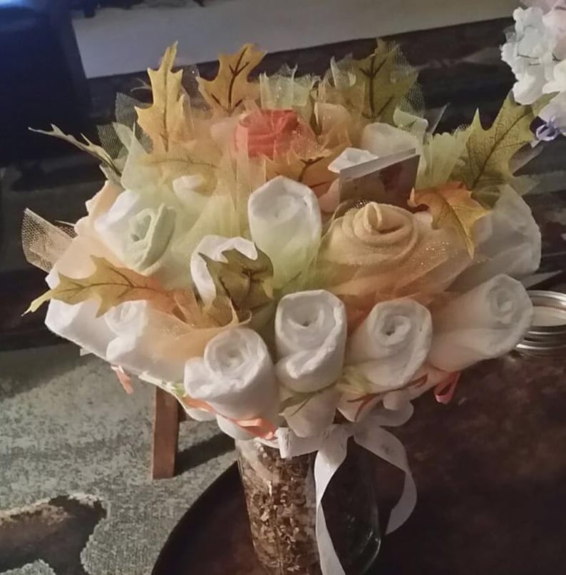 K&ouml;nnte beinhalten: Ein Strau&szlig; aus wei&szlig;en, orangefarbenen und gelben Stoffblumen und Bl&auml;ttern, angeordnet in einem durchsichtigen Glas. Die Blumen sind wie Rosen geformt und von T&uuml;ll und dekorativen Bl&auml;ttern umgeben. Ein wei&szlig;es Band ist um das Glas gebunden.