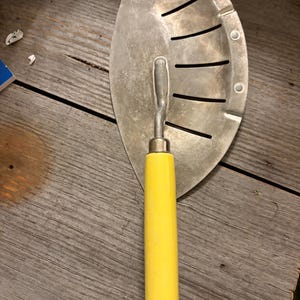 May include: A metal kitchen tool with a semi-circular, perforated blade and a bright yellow handle. The blade features a series of parallel, curved slots for draining liquids, and a small hole at the top for hanging. The tool has a sturdy, utilitarian design.